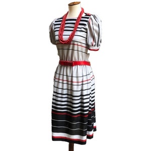 Puede incluir: Un vestido de rayas blancas y negras con un cinturón rojo y un collar de cuentas rojas. El vestido tiene mangas cortas abullonadas y una falda hasta la rodilla.