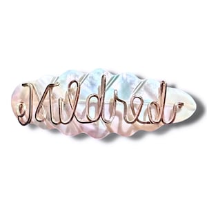 Peut inclure: Une barrette à cheveux nacrée avec le nom "Mildred" dans un design filaire délicat et cursif. La barrette a une forme ondulée en forme de nuage et un éclat irisé subtil, reflétant des nuances de rose, de bleu et de blanc.