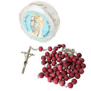 Pode incluir: Um rosário com contas vermelhas escuras e um crucifixo prateado. Também está presente um recipiente de plástico transparente com um rótulo azul com uma imagem e texto. O rótulo diz "Mare de Déu de Montserrat".