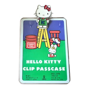 Puede incluir: Llavero con clip de Hello Kitty con una colorida ilustración de Hello Kitty, una escalera, un cubo rojo con la etiqueta "Fancy Pail", una silla roja y un conejito. El llavero tiene un borde blanco y una figura de Hello Kitty en la parte superior.