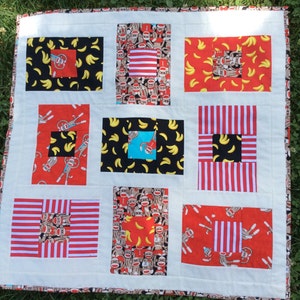 May include: A white quilt with a patchwork design featuring various fabrics with monkeys, bananas, and red and white stripes. The quilt is made up of nine squares, each with a different pattern.