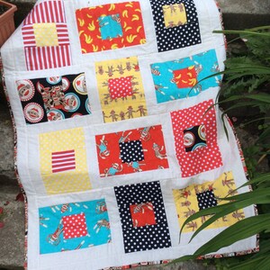 May include: A white quilt with a patchwork design featuring various colorful fabrics. The quilt is made up of squares with different patterns, including stripes, polka dots, and monkeys. The quilt is edged with a white border.