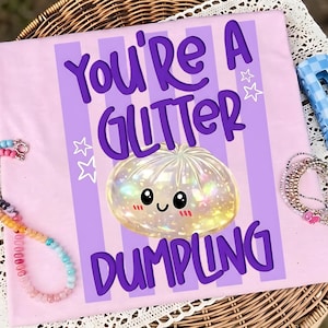May include: A light pink t-shirt featuring vertical purple stripes and the text "YOU'RE A GLITTER DUMPLING" in dark purple letters. The design includes a central illustration of a round, iridescent, glowing dumpling character with a smiling face.