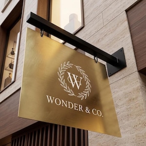 May include: A gold-colored, rectangular sign with the text "WONDER & CO." in white, hanging from a black metal bracket. The sign features a white floral wreath design around the letter "W". The sign is mounted on a stone wall.