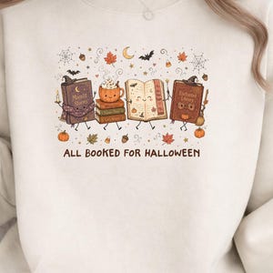 May include: A beige crewneck sweatshirt featuring a graphic print of anthropomorphic books, a pumpkin mug, and autumn elements like leaves, bats, and spiderwebs. The text below the illustration reads ALL BOOKED FOR HALLOWEEN in brown letters.