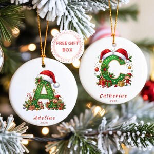 May include: Two round white ceramic ornaments hanging by gold strings. One features a green glittery letter A with a Santa hat, an elf, and gifts, labeled Adeline 2026. The other features a green glittery letter C, an elf, and gifts, labeled Catherine 2026.