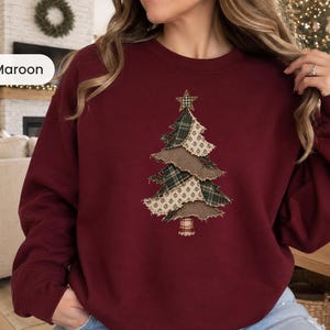 May include: A maroon crewneck sweatshirt featuring a central graphic of a Christmas tree constructed from layered, frayed fabric patches in green plaid and cream-colored patterns, topped with a small star and a plaid trunk.