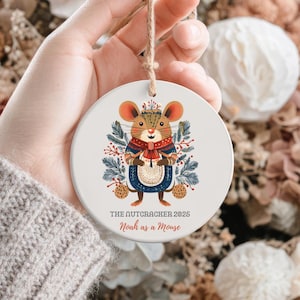 Può includere: Ornamento natalizio rotondo in ceramica bianca con un disegno di topo dei cartoni animati. Il topo indossa un abito blu e rosso ed è circondato da foglie. Il testo sull'ornamento recita "THE NUTCRACKER 2025 Noah as a Mouse."