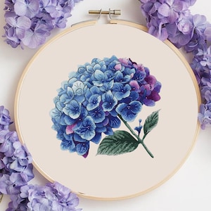 Puede incluir: Bordado de punto de cruz de una flor de hortensia azul y morada. La flor está centrada en una tela color crema, enmarcada por un aro de bordar de madera. Se ven hojas y tallos verdes. Flores moradas alrededor del aro.