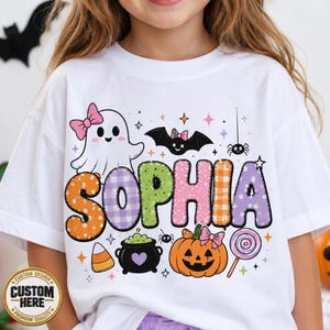 May include: A white t-shirt featuring the name SOPHIA in large, colorful, patterned letters. The design includes a ghost with a pink bow, a bat, a spider, a black cauldron with a purple heart, a jack-o'-lantern with a pink bow, and a lollipop.