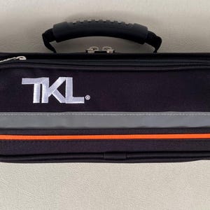 May include: Black rectangular case with a handle and a zippered pocket. The case has a reflective gray and orange stripe design. The text "TKL." is printed in white on the front of the case.