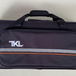 May include: Black rectangular instrument case with a zippered front pocket. The pocket has a reflective gray stripe with an orange accent. The text "TKL" is printed in white on the pocket. A black handle is on top.