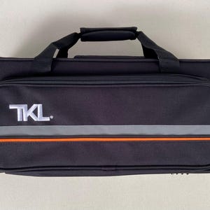 May include: Black rectangular carrying case with a handle and a zippered front pocket. The front pocket has the text "TKL" in white, with a gray and orange stripe design.