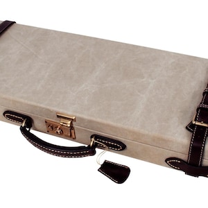 May include: A rectangular, light gray case with dark brown leather accents. The case features two leather straps with buckles, a handle, and a lock. The corners are reinforced with dark brown leather, and a small tag hangs from the handle.