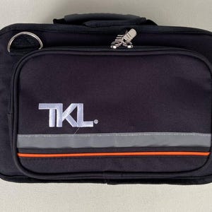 May include: Black rectangular bag with a handle and a shoulder strap ring. The bag has a front pocket with the white letters "TKL" and a reflective gray and orange stripe. A silver zipper is visible.