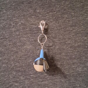 Peut inclure: Un porte-cl&eacute;s avec un mousqueton et un anneau argent&eacute;s, repr&eacute;sentant une petite figurine stylis&eacute;e. La figurine a une t&ecirc;te bleue, un corps beige et des d&eacute;tails noirs. Le porte-cl&eacute;s est suspendu sur un fond gris textur&eacute;.