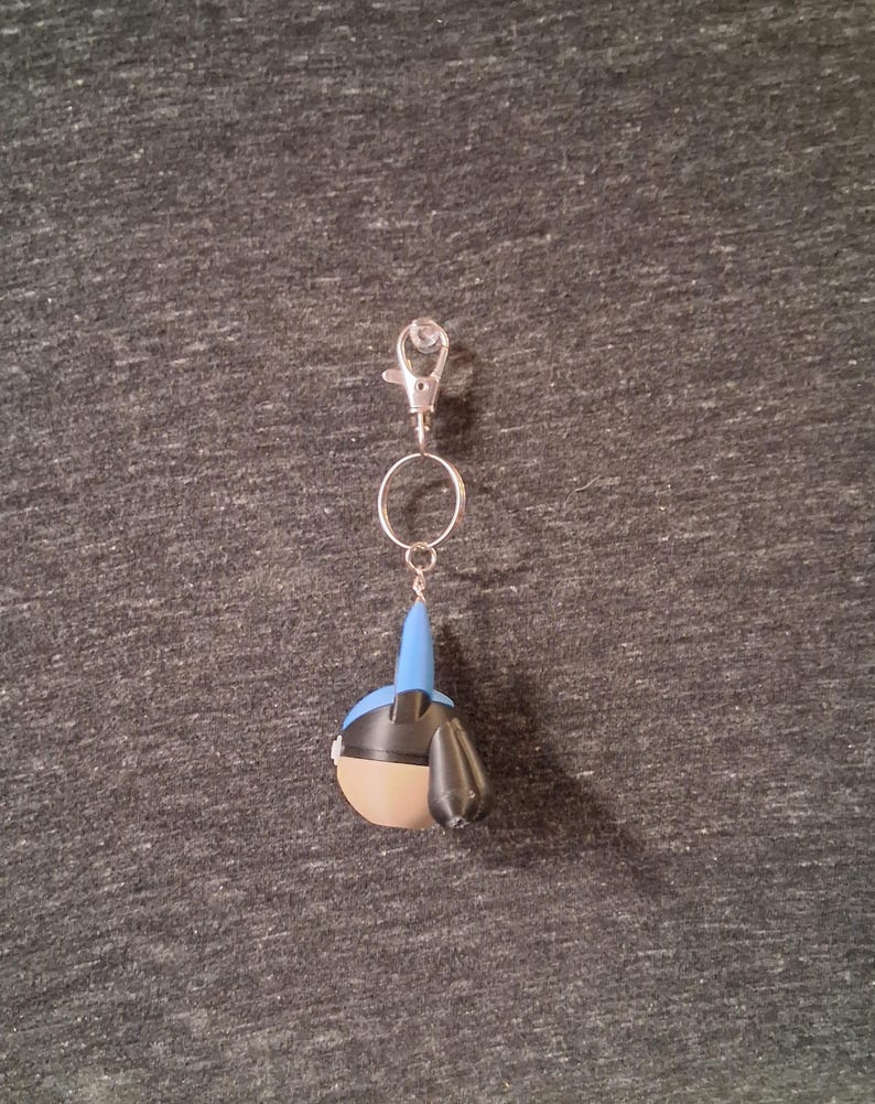 Peut inclure: Un porte-cl&eacute;s avec un mousqueton et un anneau argent&eacute;s, repr&eacute;sentant une petite figurine stylis&eacute;e. La figurine a une t&ecirc;te bleue, un corps beige et des d&eacute;tails noirs. Le porte-cl&eacute;s est suspendu sur un fond gris textur&eacute;.