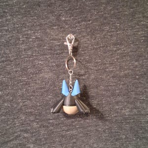 Peut inclure: Un porte-cl&eacute;s avec une figurine noire et bleue, un fermoir argent&eacute; et une cha&icirc;ne. La figurine a des oreilles pointues bleues et un ventre beige. Le porte-cl&eacute;s est attach&eacute; &agrave; un fermoir en m&eacute;tal argent&eacute;.