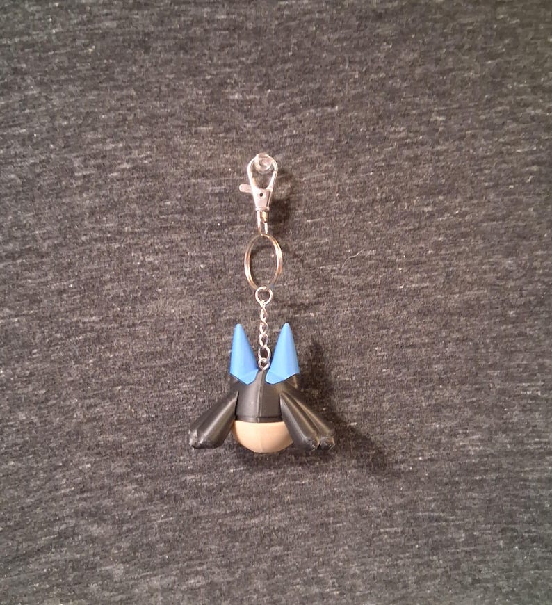 Peut inclure: Un porte-cl&eacute;s avec une figurine noire et bleue, un fermoir argent&eacute; et une cha&icirc;ne. La figurine a des oreilles pointues bleues et un ventre beige. Le porte-cl&eacute;s est attach&eacute; &agrave; un fermoir en m&eacute;tal argent&eacute;.
