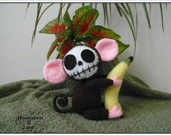 May include: A knitted toy monkey with a skull face and pink ears holds a yellow knitted banana. The monkey is sitting on a green towel.