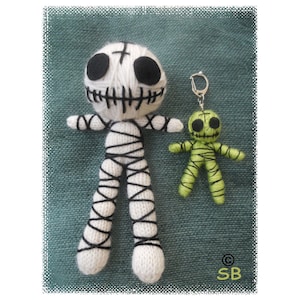May include: Two crocheted voodoo dolls, one white with black stitching and a cross on its forehead, and one green with black stitching. The larger doll is standing on a blue fabric background, while the smaller doll is attached to a silver key chain.