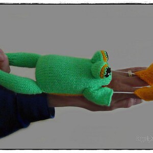 May include: A green and orange knitted frog puppet with a hand inside. The frog has large eyes and a wide mouth. The puppet is being held up by a hand.