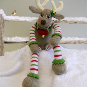 May include: A knitted reindeer toy with red and white striped stockings, green and white striped sleeves, and a red bow. The reindeer is sitting on a white fluffy surface with a small red bird on a wooden fence in the background.