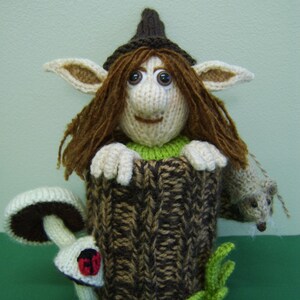 May include: A knitted elf doll with brown hair and a pointed hat is peeking out of a brown and white knitted tree stump. The elf is holding a small knitted mouse in its hand. There is a white mushroom with red spots next to the stump.
