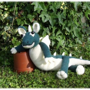 May include: A handcrafted knitted dragon toy in teal and cream, leaning on a brown pot. The dragon features white wings, ears, and a tail, set against a backdrop of green foliage and grass.