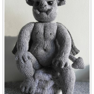 Può includere: Una scultura di gargoyle di lana grigia seduta su una superficie grigia. Il gargoyle ha un viso dettagliato con occhi grandi e una bocca larga. La scultura è realizzata in lana e ha una superficie testurizzata.