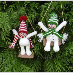May include: Two knitted snowmen wearing hats and scarves are swinging on a rope swing. The snowmen are white with black eyes and mouths. The snowman on the left is wearing a red and white striped hat and scarf. The snowman on the right is wearing a green and white striped hat and scarf. The snowmen are hanging from a tree branch.