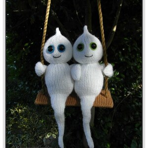 Op de afbeelding: Twee witte gebreide spookfiguren met blauwe en groene ogen zitten op een bruine schommel tegen een groene, bladerrijke achtergrond.