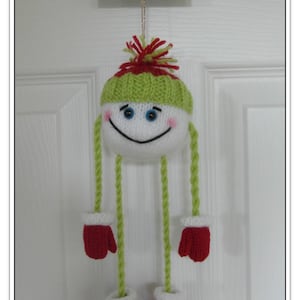 May include: A knitted snowman ornament with a green hat, red mittens, and a white face. The ornament is hanging from a white door with a sign that says "Happy Holidays".