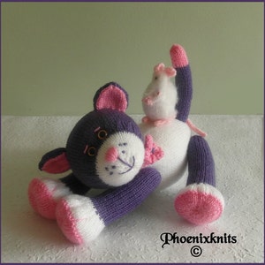May include: A purple and white knitted cat toy with pink accents and a small white knitted mouse toy on its back. The cat toy has a pink nose and black eyes.