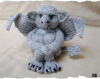 Op de afbeelding: Een grijze gebreide gargoyle met een humeurige uitdrukking, zittend met gekruiste armen en een staart die achter hem is opgerold.