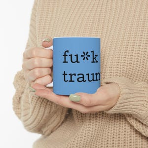 Könnte beinhalten: Eine hellblaue Keramiktasse mit den Worten "fu*k traum" in Schwarz aufgedruckt. Die Tasse wird von einer Person gehalten, die einen beigen Pullover trägt. Die Tasse hat einen weißen Innenraum.