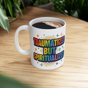 May include: White ceramic mug with the text "TRAUMATIZED BUT SPIRITUALIZED" in rainbow colors. The mug is filled with dark coffee and sits on a wooden surface. The design includes stars and a rainbow.