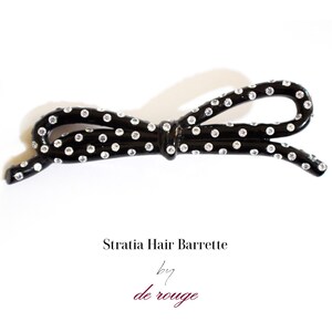 Puede incluir: Barreta para el pelo negra con lunares blancos y pedrería. La barrita tiene forma de lazo. El texto "Stratia Hair Barrette by de rouge" está impreso debajo de la barrita.