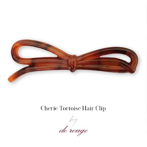 Peut inclure: Une pince à cheveux en forme de nœud en écaille de tortue brune. La pince est en plastique et a une finition brillante. Le texte "Cherie Tortoise Hair Clip by de rouge" est imprimé sous la pince.