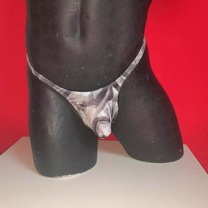 Könnte beinhalten: Ein grau-weiß gemusterter String-Tanga auf einem schwarzen Mannequin-Torso. Der String hat einen dünnen Bund und einen Beutel. Der Hintergrund ist rot und die Mannequin steht auf einer weißen Oberfläche.