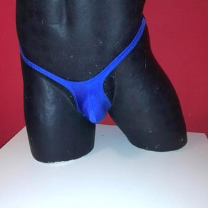Può includere: Un perizoma blu esposto su un manichino nero. Il costume da bagno ha un design semplice con spalline sottili e un davanti triangolare. Lo sfondo è di colore rosso pieno e il manichino è su una superficie bianca.