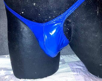 May include: A shiny, royal blue thong swimsuit bottom. The fabric has a glossy appearance. The garment is displayed on a black mannequin. The background is a white and gray patterned surface.