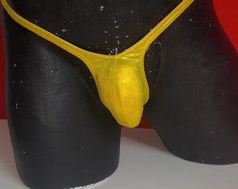 May include: A gold metallic thong swimsuit bottom. The garment is displayed on a black mannequin. The background is red and white. The swimsuit bottom is a solid color and has a shiny appearance.