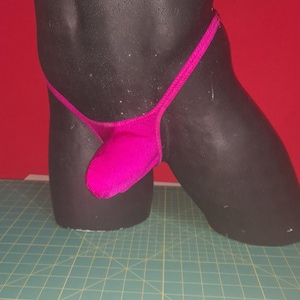 Könnte beinhalten: Ein magentafarbener String-Badeanzug, der auf einer schwarzen Schaufensterpuppe ausgestellt ist. Der Badeanzug hat ein minimalistisches Design mit einem dünnen Bund und einem Beutel. Der Hintergrund ist einfarbig rot, und die Schaufensterpuppe steht auf einer grünen Schneidematte.