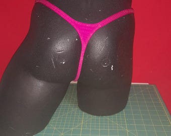 May include: A hot pink thong swimsuit bottom displayed on a black mannequin. The thong has a thin waistband and string sides. The mannequin is on a green cutting mat with a red background.
