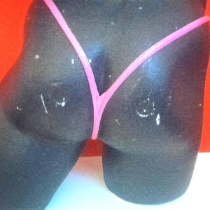 May include: A black swimsuit bottom with a thin pink strap design. The garment is displayed on a mannequin against a bright red background. The bottom is a thong style.