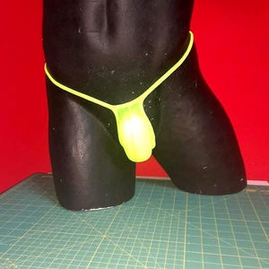 Può includere: Un perizoma verde neon su un manichino nero. La biancheria intima ha un sottile cordino giallo brillante e una tasca. Lo sfondo è rosso.