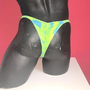 May include: A mannequin torso is shown from the back, wearing a tie-dye bikini bottom. The bikini bottom is a vibrant mix of yellow, blue, and green. The background is a solid red color, and the mannequin is on a white surface.
