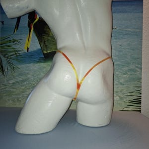 Puede incluir: Un torso de maniquí blanco que lleva un traje de baño tanga con ribete naranja y amarillo. El maniquí está colocado frente a un telón de fondo de escena de playa. El traje de baño es el punto focal, destacando la forma.