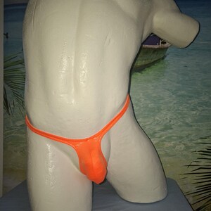 Könnte beinhalten: Ein orangefarbener String-Badeanzug, der auf einer weißen Schaufensterpuppe präsentiert wird. Der Badeanzug hat einen dünnen orangefarbenen Riemen und einen Beutel. Die Schaufensterpuppe befindet sich vor einem Strandhintergrund.